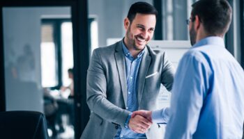 two-smiling-businessmen-shaking-hands-while-standing-office-min